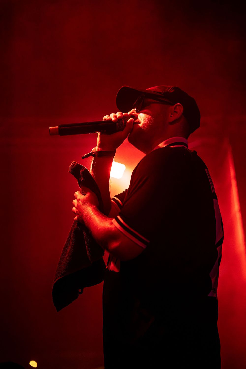 A male rapper performs on stage under dramatic red lighting, holding a microphone close to his face while wearing a black cap.