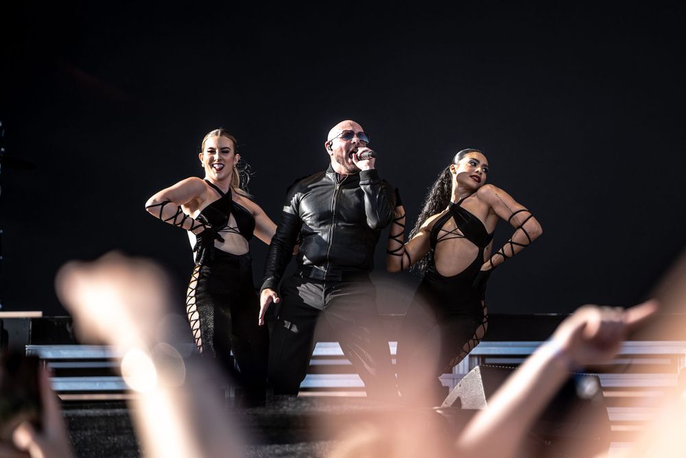 Male performer in black leather jacket sings on stage flanked by two female dancers in black outfits under dramatic lighting.
