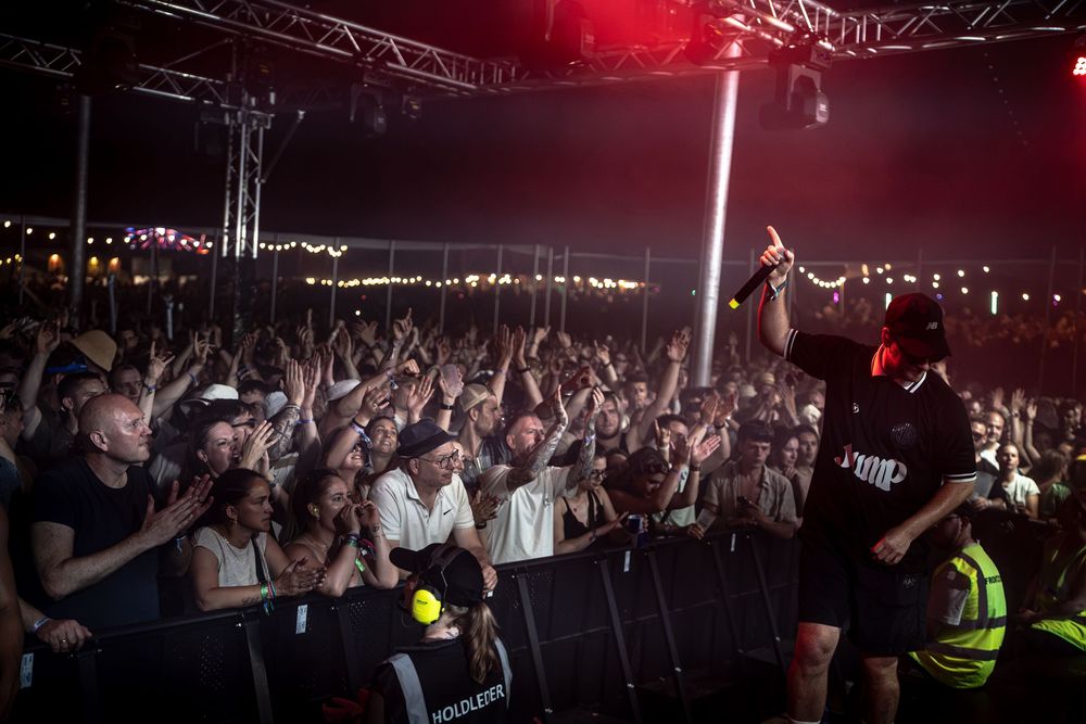 Performer on stage raises microphone to energetic crowd at outdoor music festival at night.