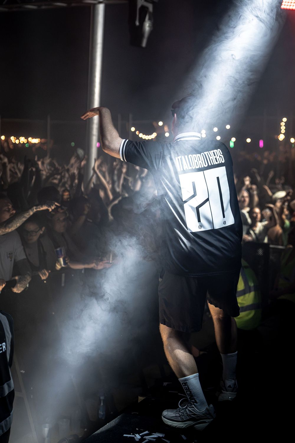 Performer in black jersey number 20 faces crowd on smoky concert stage with dramatic spotlight.