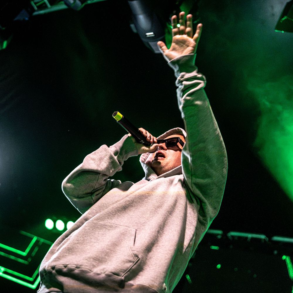 A performer in a white hoodie and sunglasses holds a microphone while raising one hand on a green-lit concert stage.