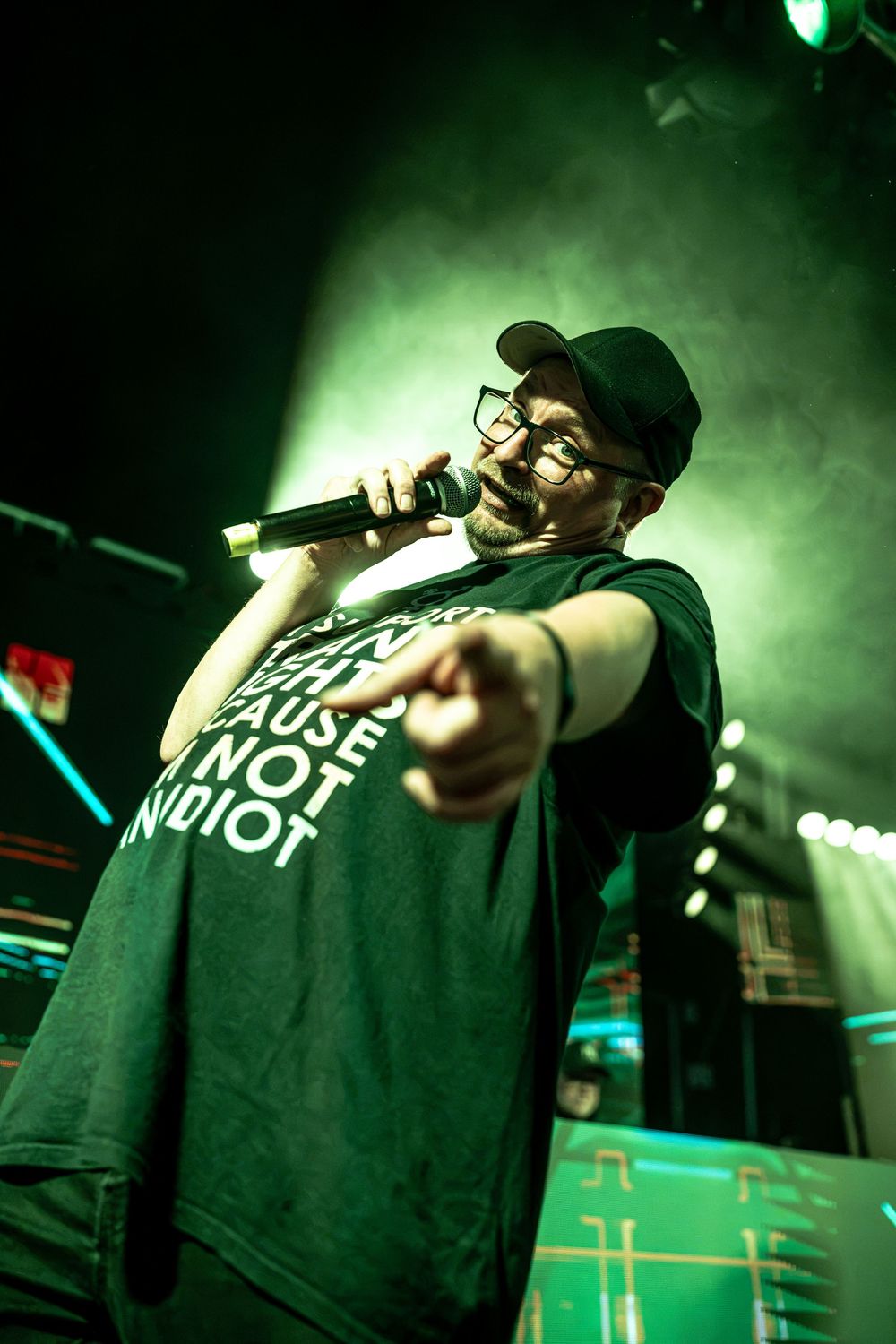 A performer in a black cap and glasses sings into a microphone on a green-lit stage, pointing at the crowd.
