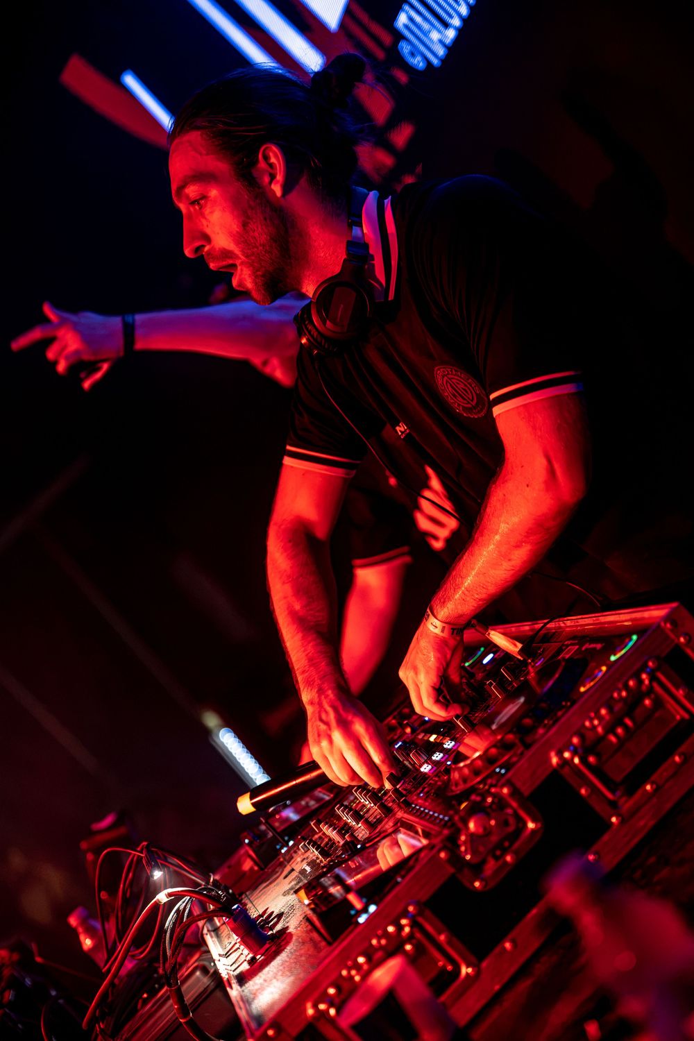 A DJ performing at a nightclub, leaning over mixing equipment under dramatic red lighting with headphones around his neck.