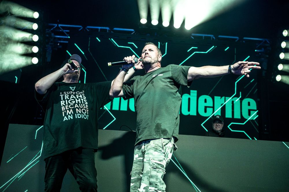 Two rappers performing on a neon-lit stage, one wearing a trans rights shirt, microphones in hand.