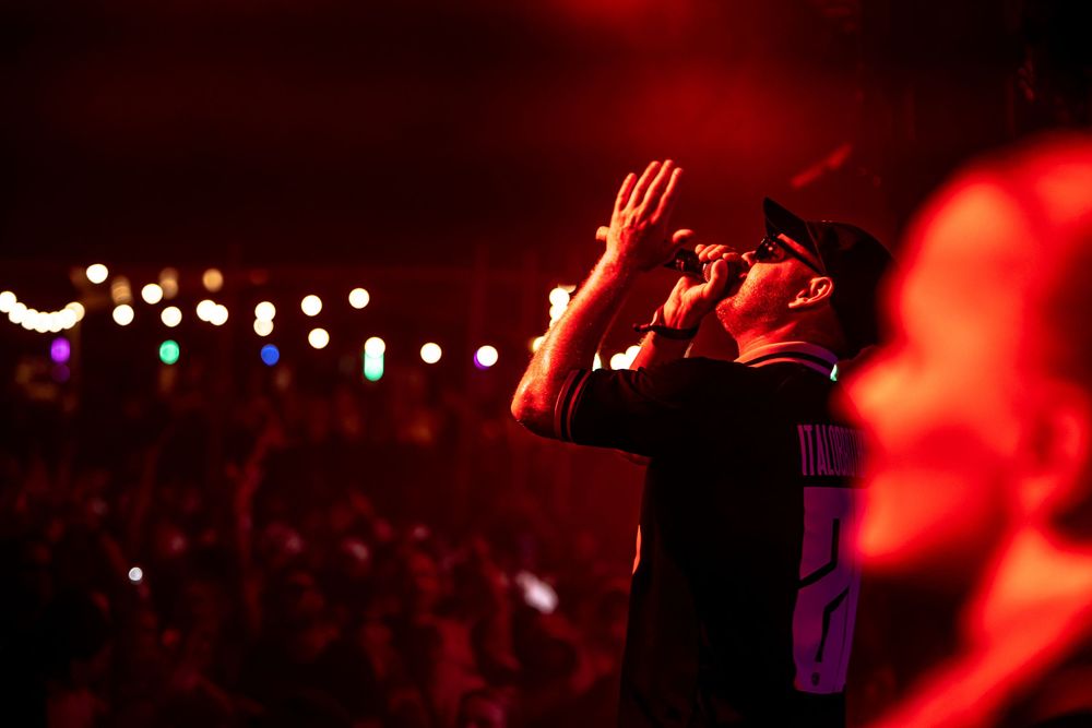 Rapper performing on stage with microphone raised hand crowd lit by red lights at outdoor music festival.