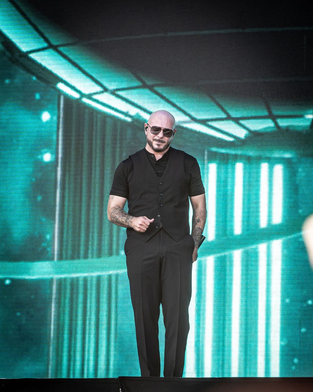 Bald man in black vest and pants smiling on stage with teal LED lights backdrop at live performance.