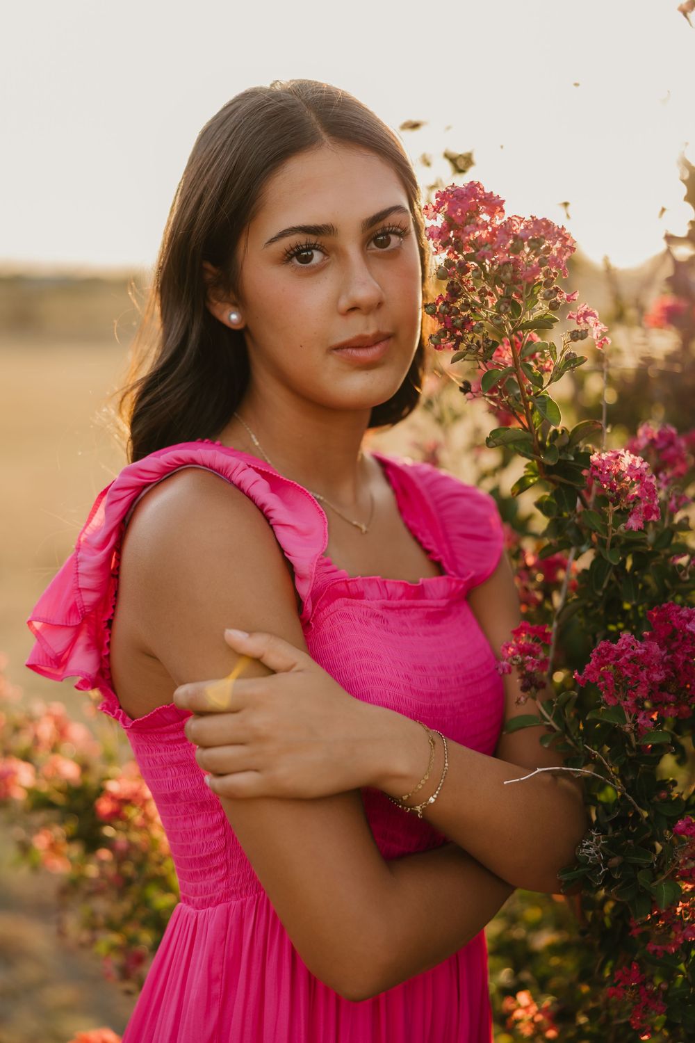 Senior poses with a serious face for portraits in Tulia Texas in front of pink flowers wearing a matching pink dress during golden hour