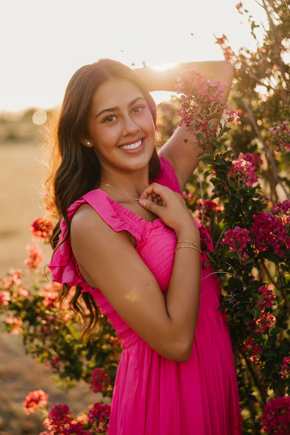 A brunette girl wearing pink dress smiles with sunlight streaming behind her as she poses for senior portraits in Tulia Texas