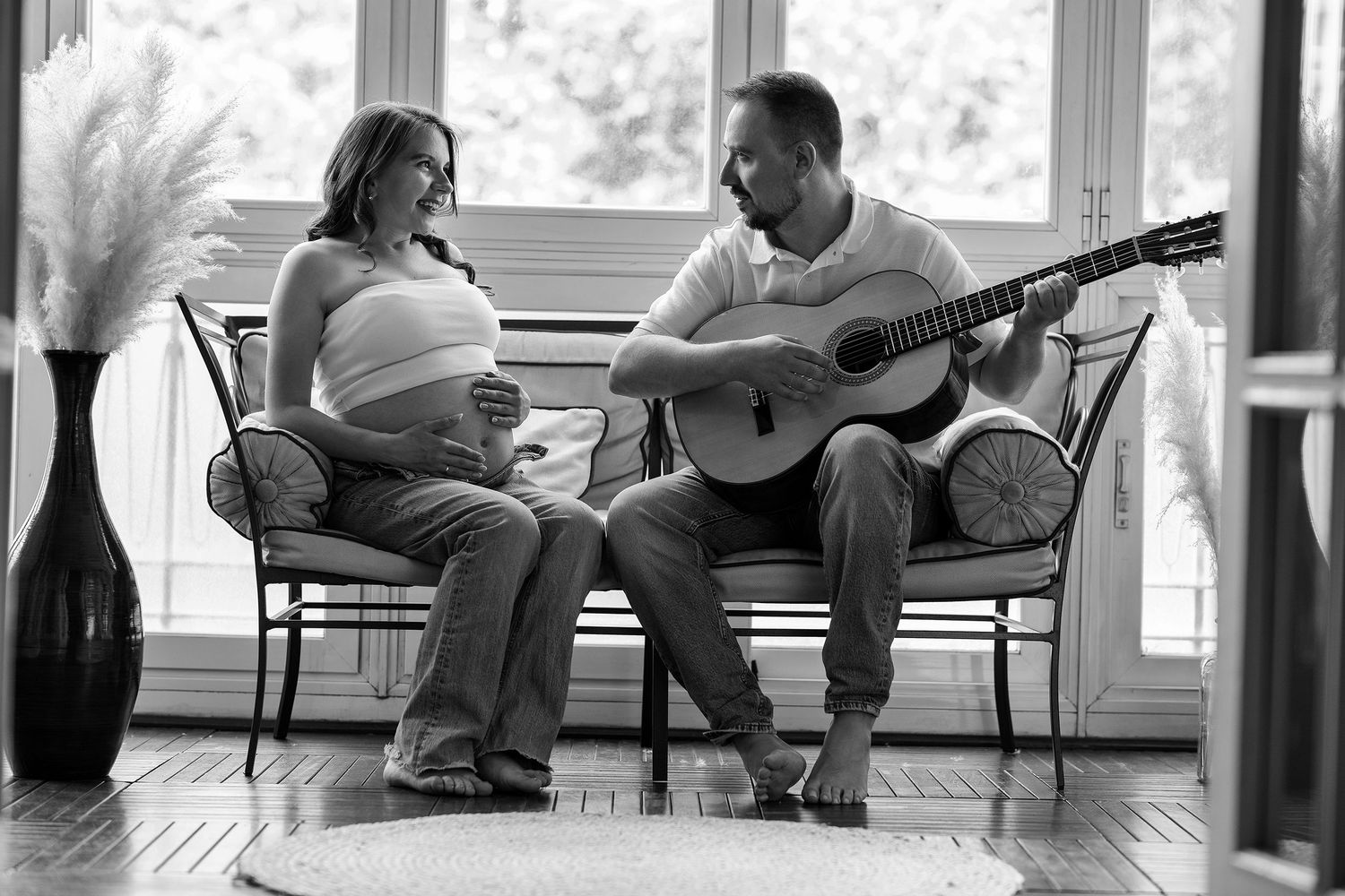 Black and white photo of pregnant woman and man playing acoustic guitar together in sunlit room.