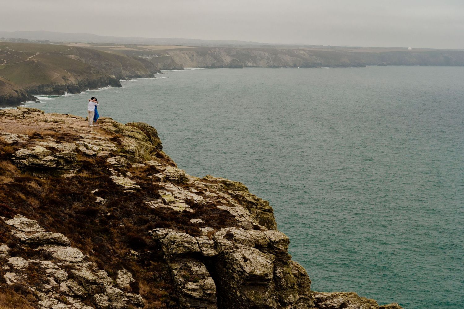 A couple stands on the cliff at st agnes head overlooking a misty turquoise coastline with rolling hills in the background.