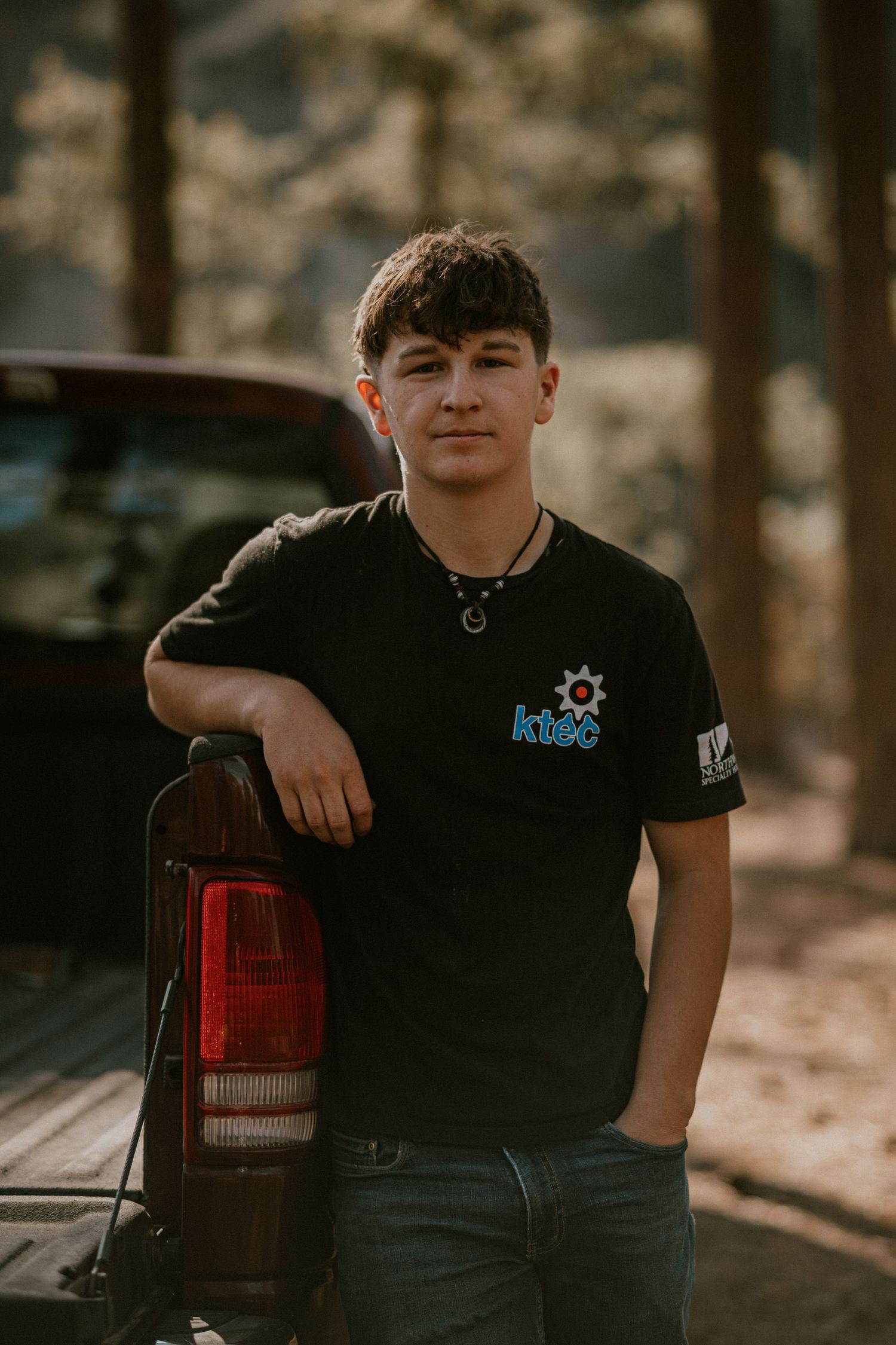 Young man in black KBEC t-shirt leaning on red truck tailgate outdoors in wooded area.