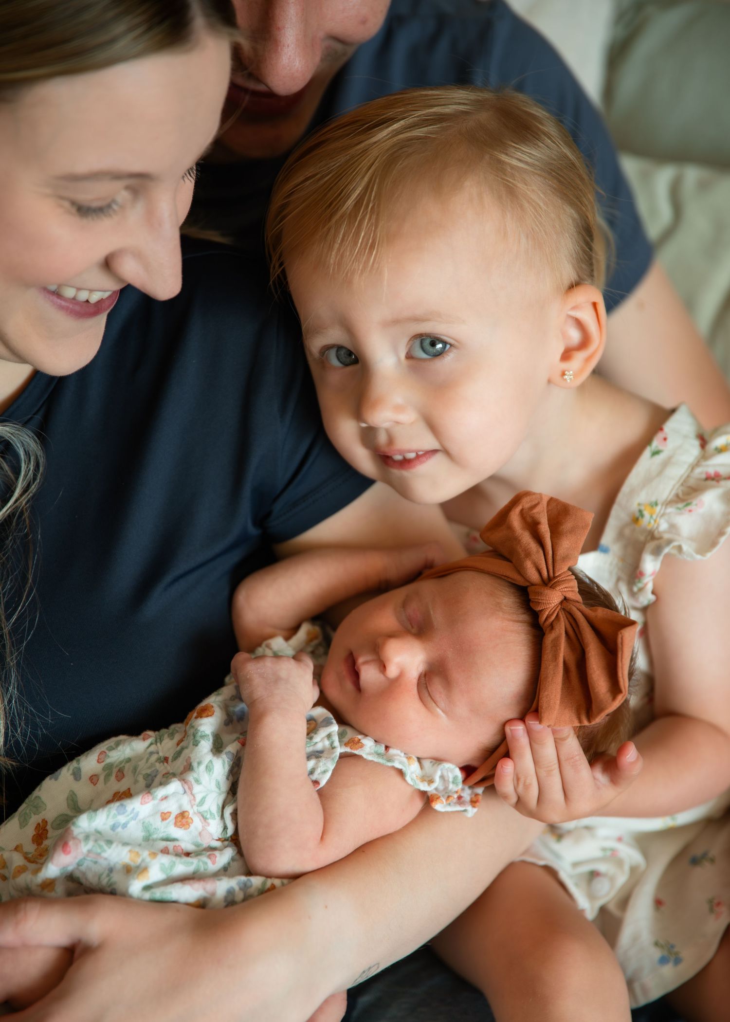 A family tenderly gazes at their sleeping newborn baby, held by a smiling toddler girl in a floral dress.