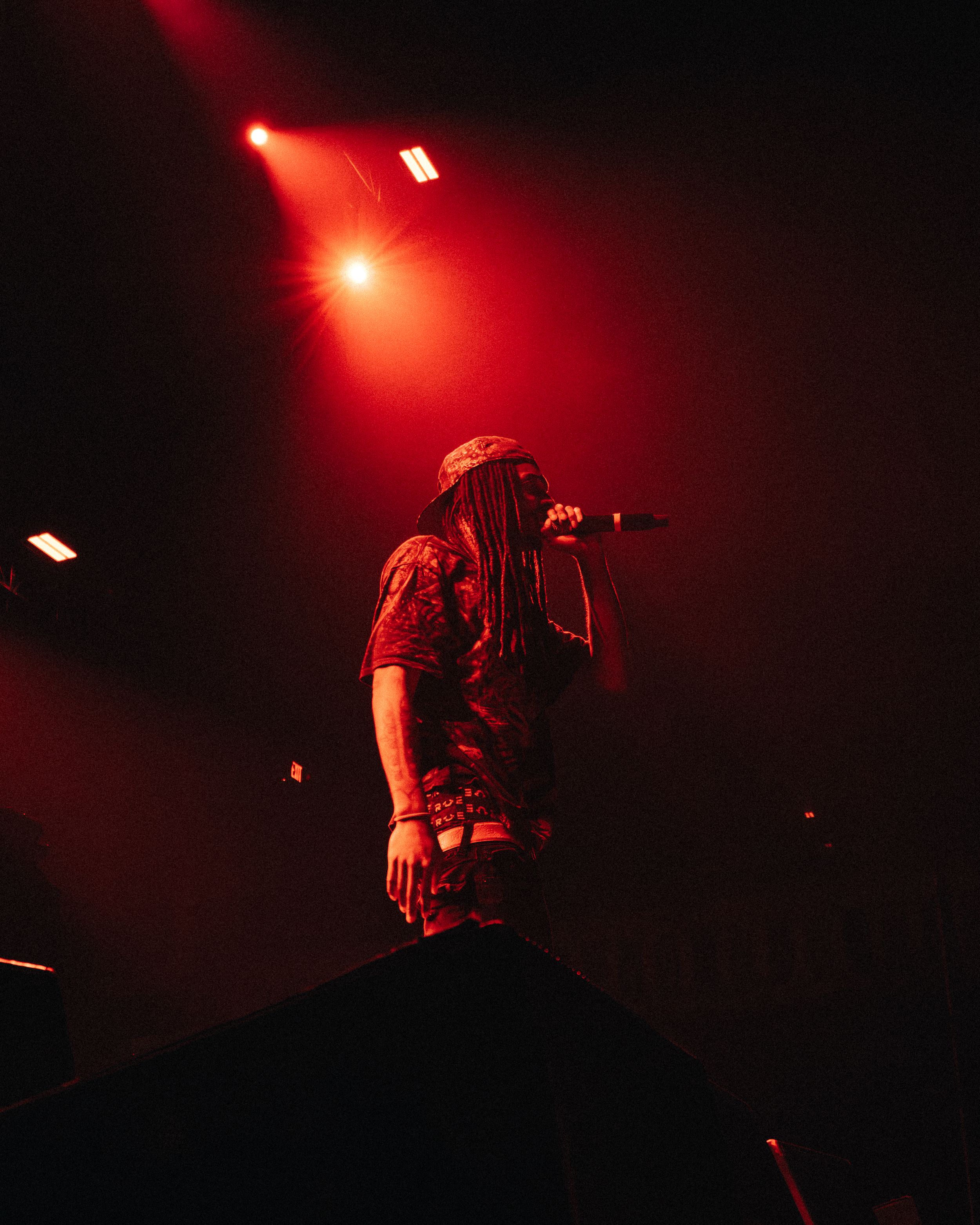 A performer with dreadlocks sings into a microphone on stage under dramatic red lighting at a live concert.