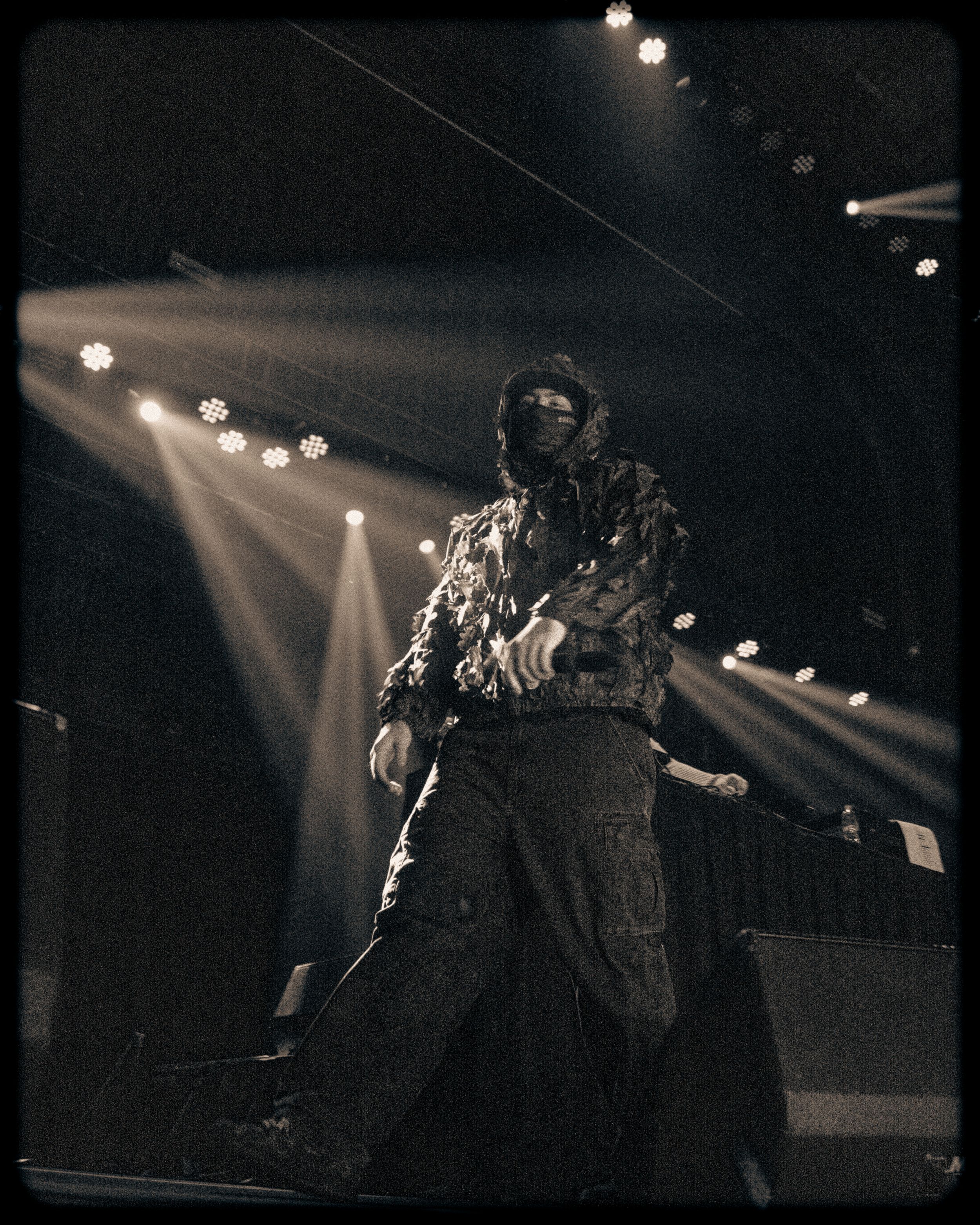 Performer in costume on stage with dramatic beam lighting in sepia-toned concert photo.