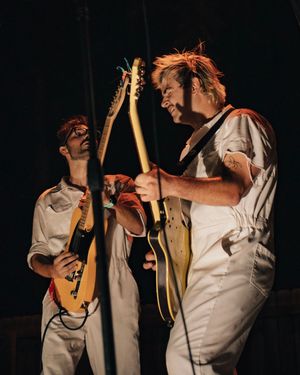 Two musicians in white overalls perform on stage, one playing electric guitar and another holding a lute-like instrument.