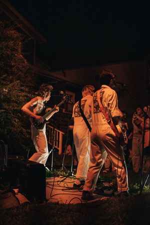 Band performing outdoors at night, musicians in white overalls playing guitar under warm ambient lighting.