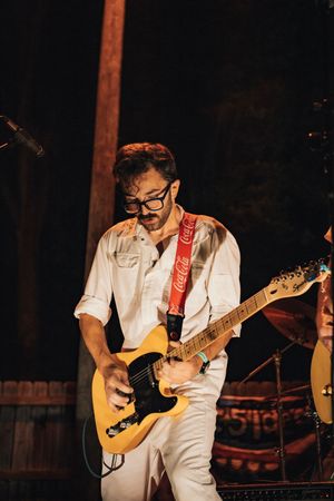 Musician in white outfit plays yellow electric guitar with Coca-Cola strap on stage under warm lights.