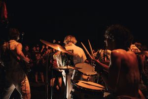 Shirtless tribal performers play drums on a dark stage before a crowd at an outdoor concert.