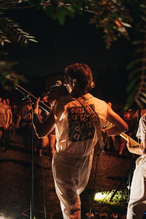 Singer performing outdoors at night holding microphone wearing white jumpsuit with graphic back print at outdoor concert.