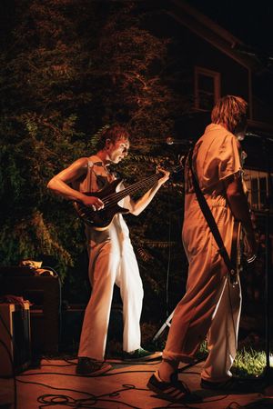Two musicians performing outdoors at night, one playing electric guitar in white jumpsuit at backyard concert.