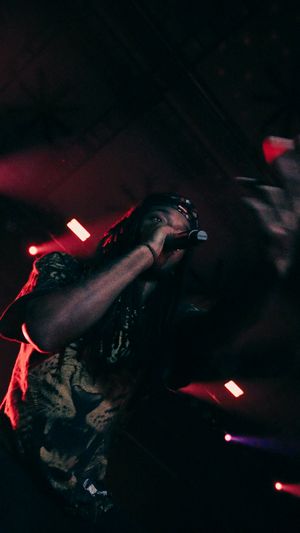 A performer passionately raps into a microphone on a dark stage illuminated by red and purple concert lights.