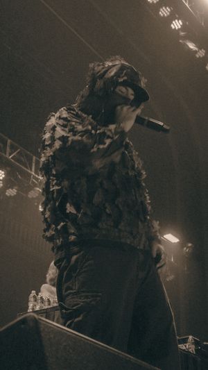 Performer in textured costume holding microphone on stage under dramatic concert lighting.
