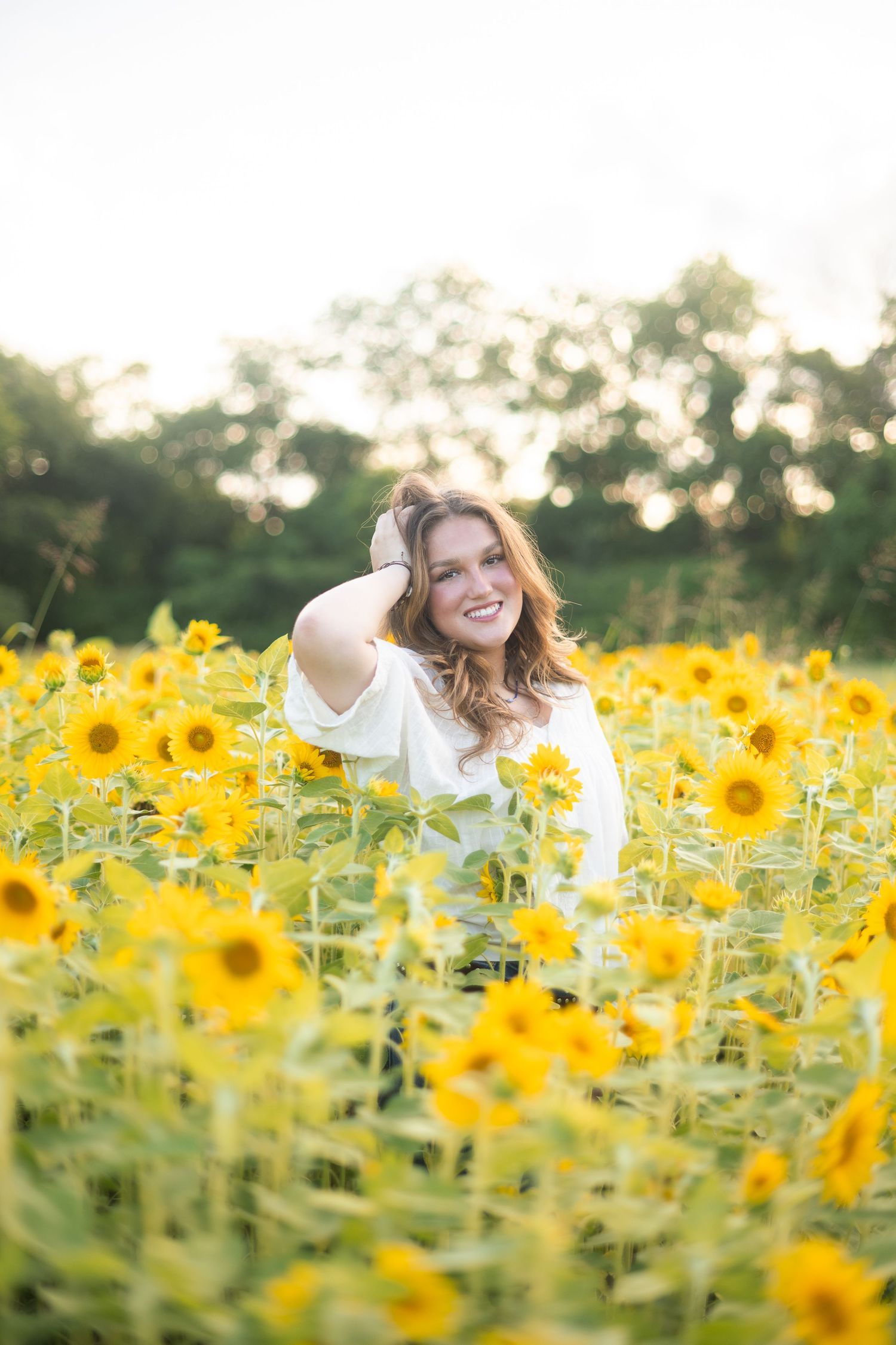 Smiling young woman in white top standing in a vibrant sunflower field with trees in the background.