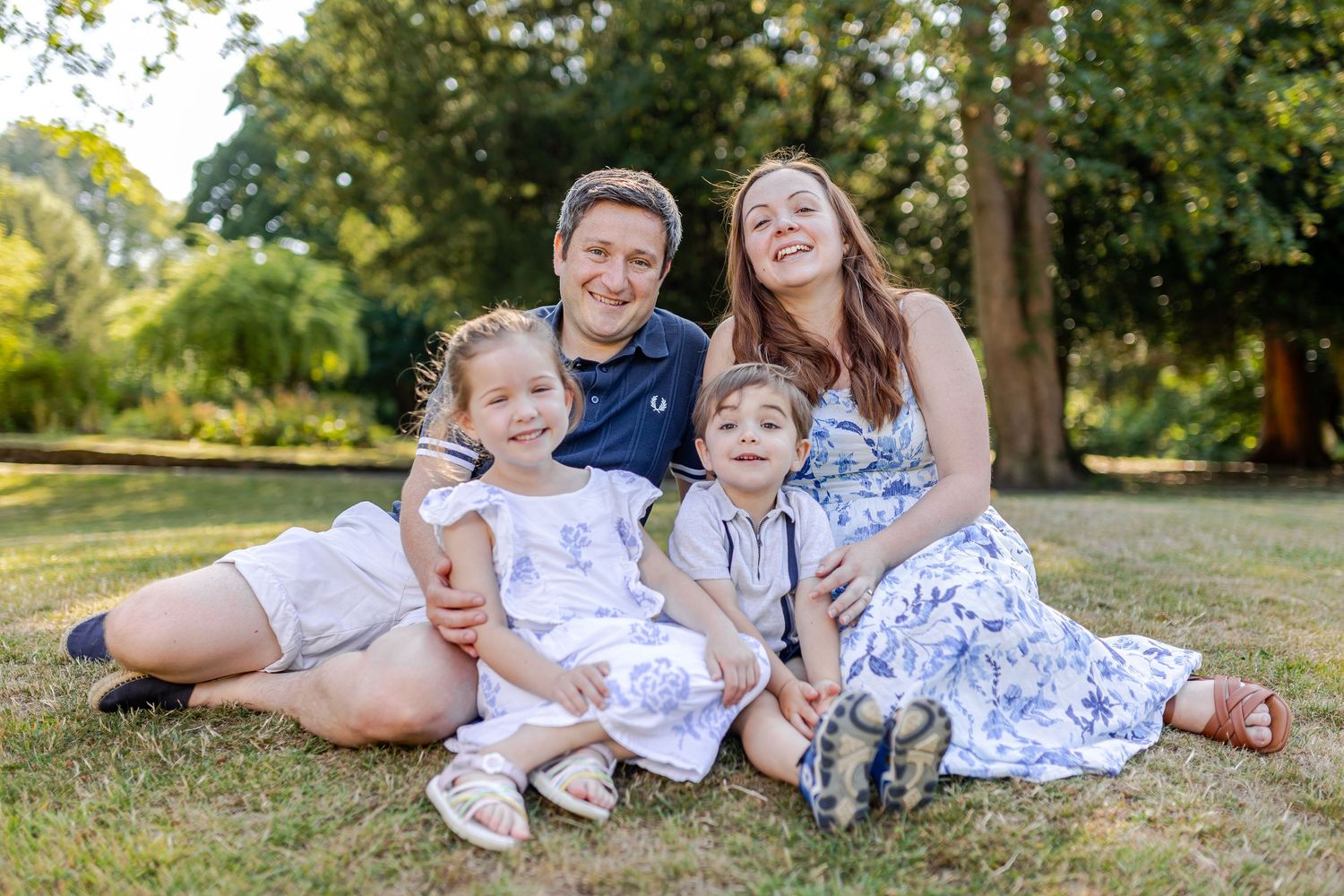 Happy family of four sitting on grass in a sunny park, parents smiling with two young children in white outfits.