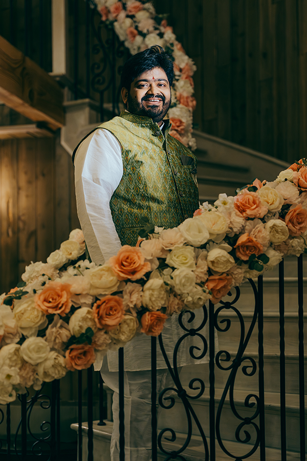 Smiling man in green embroidered vest stands on floral-decorated staircase with orange and white roses at Indian wedding.