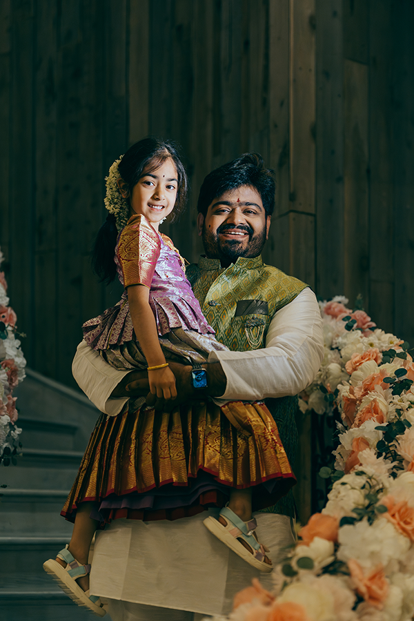 Man in traditional Indian attire holds a young girl in colorful lehenga, both smiling at a floral decorated event.