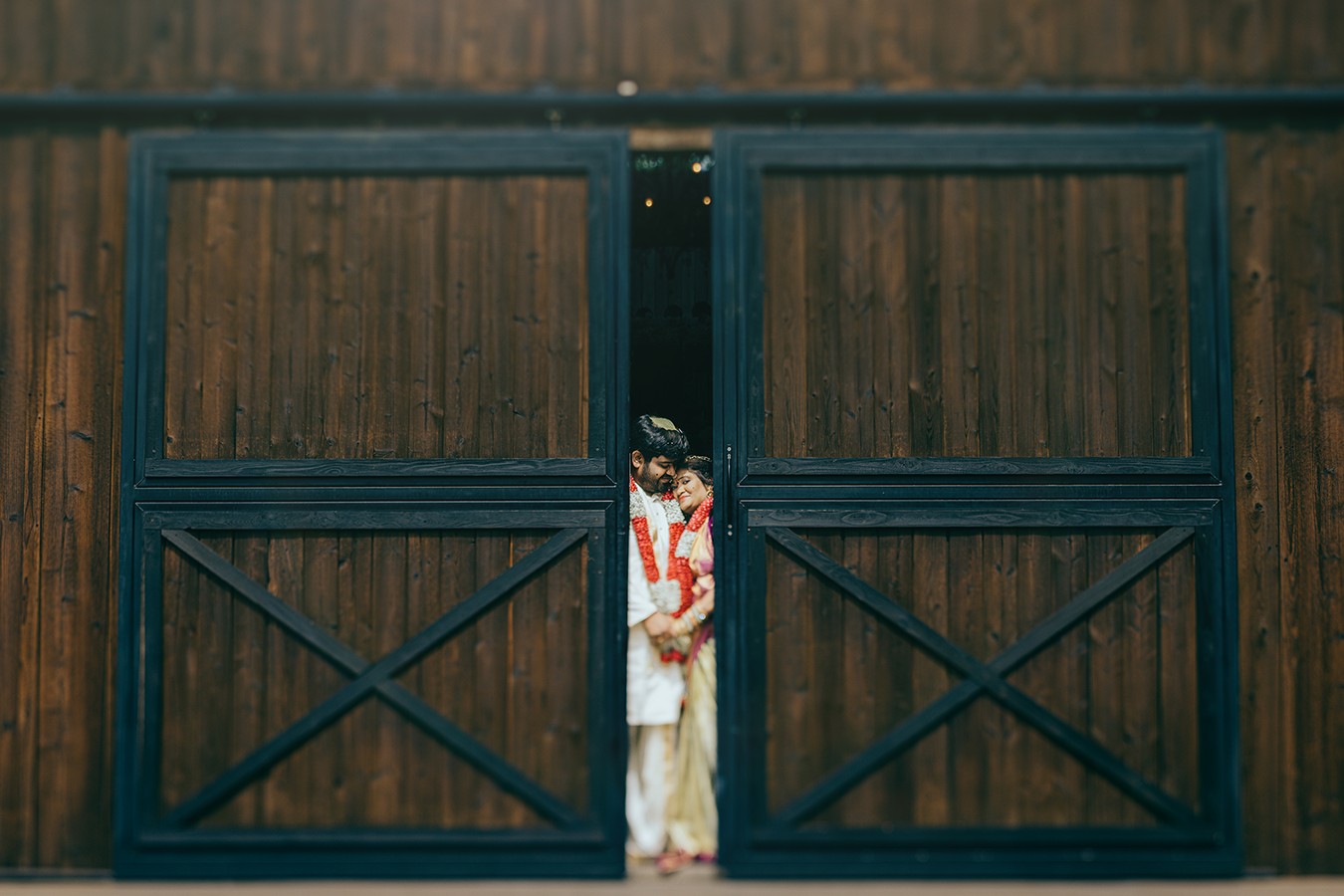 Bride in traditional attire standing between large wooden barn doors with blue trim at a rustic wedding venue.