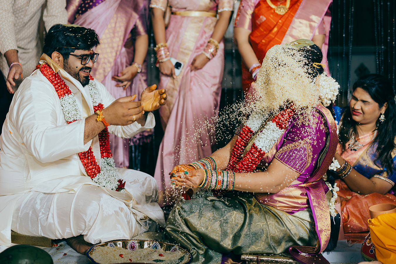 Traditional South Indian wedding ceremony with groom in white and bride in purple saree exchanging flower garlands.
