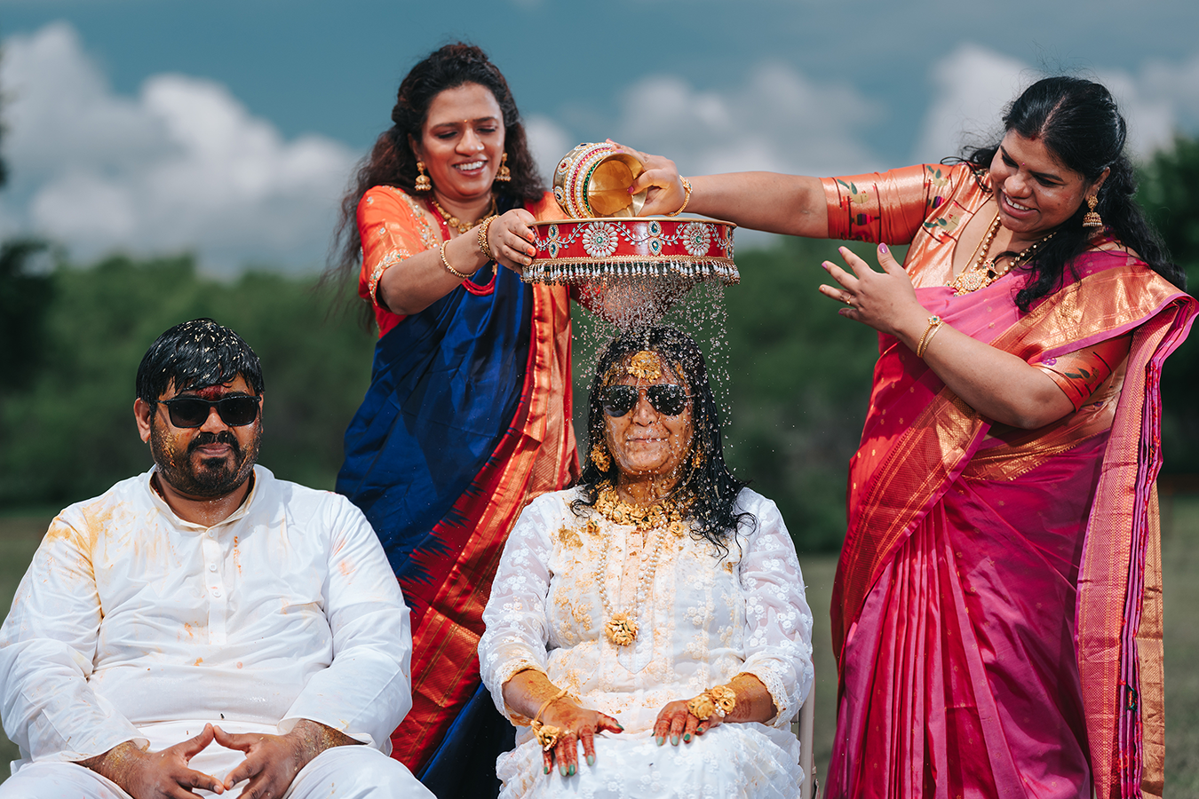 Indian bride in white outfit receives traditional haldi ceremony blessing from women in colorful silk sarees outdoors.
