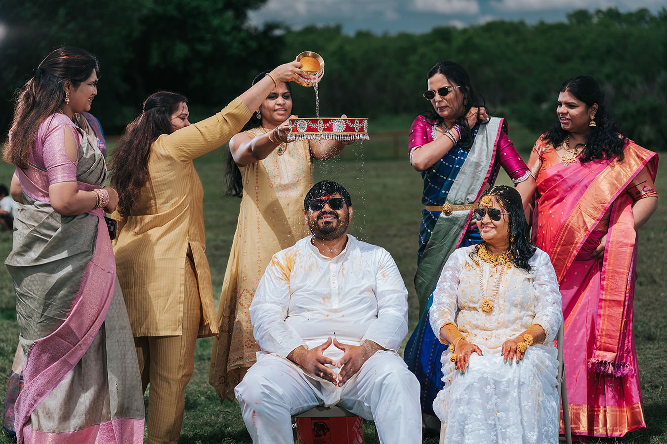 Indian wedding ceremony with bride and groom seated outdoors while women in colorful sarees perform traditional rituals.