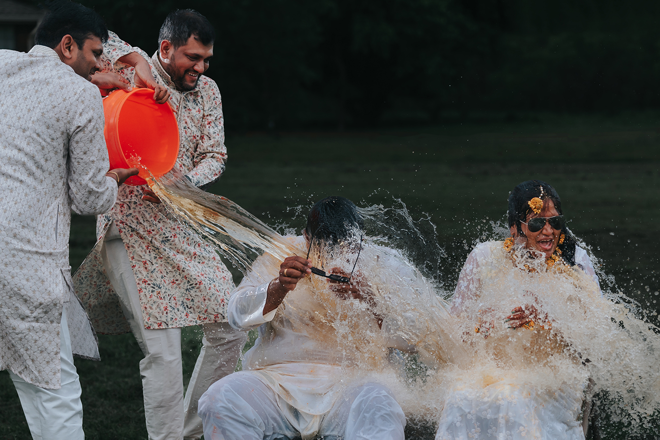 Men in white traditional attire laugh as water splashes during a festive Haldi ceremony outdoors.