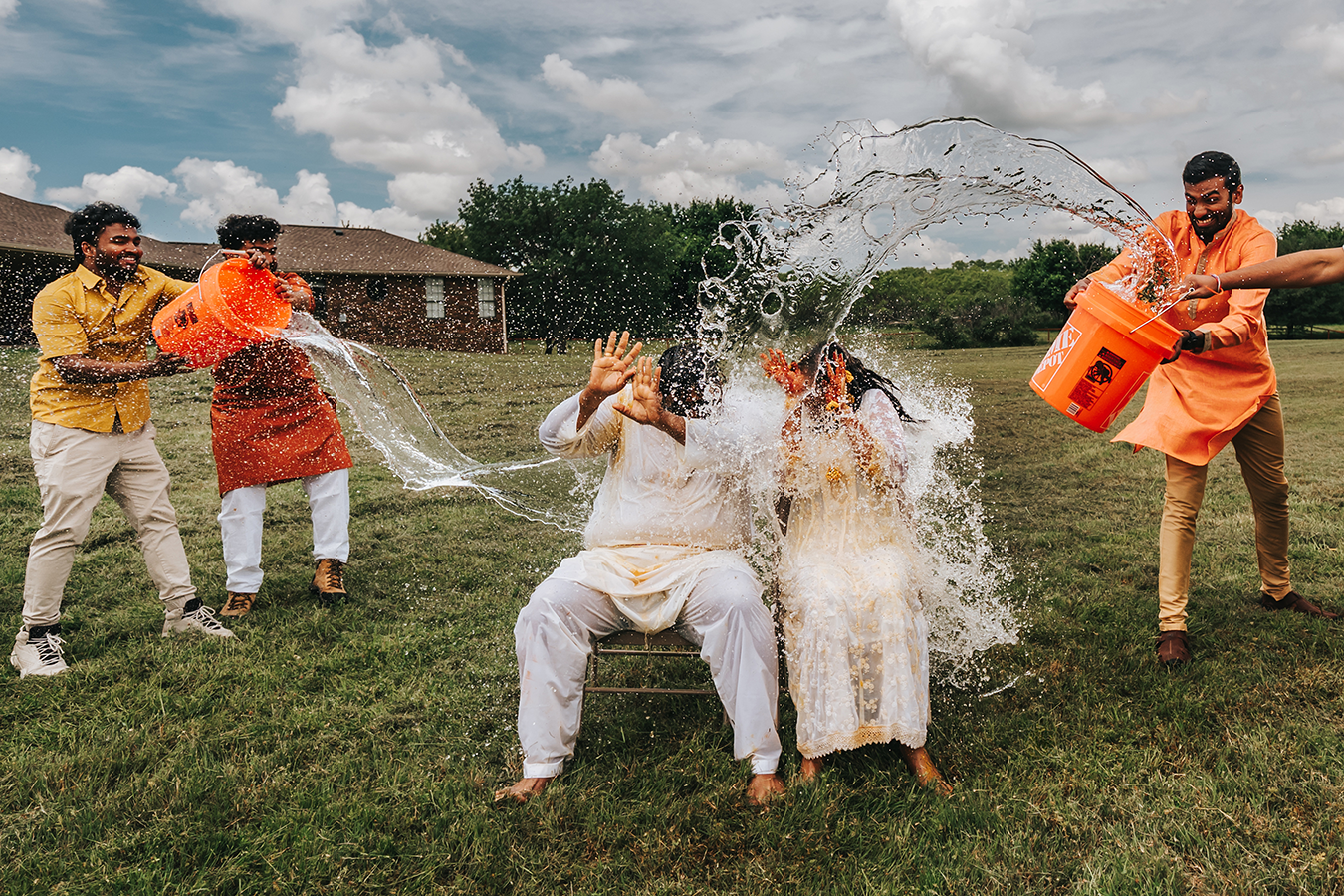 People celebrating Holi festival outdoors, joyfully splashing water on two seated individuals in white traditional Indian attire.