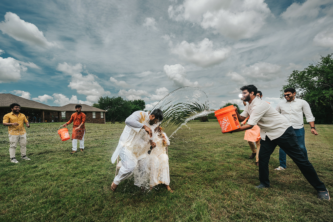 People joyfully splashing water on a couple during an outdoor Indian wedding celebration on a green lawn.