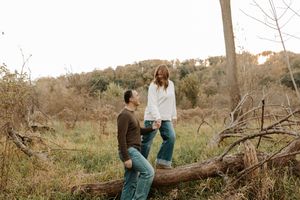 Couple in casual sweaters and jeans share a quiet moment beside a fallen log in an autumn woodland field with golden grass and bare trees.