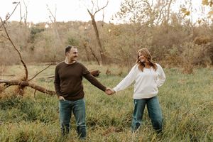 Smiling couple holding hands and walking through a grassy autumn field with bare trees and fallen branches in the background.
