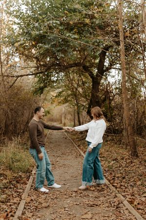 Couple holding hands and smiling on a leaf-covered wooden path in an autumn forest during an engagement photo session.