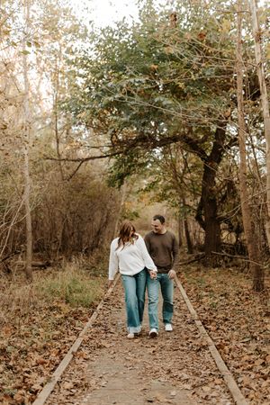 Couple in jeans walking hand in hand down a narrow wooden boardwalk surrounded by fall trees and fallen leaves.
