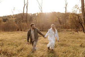 Couple holding hands and walking through a golden grassy field with bare trees and warm sunset light in the background.
