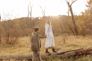 Woman in a white dress walks along a fallen log holding hands with a man in a tan jacket in a golden autumn field at sunset.