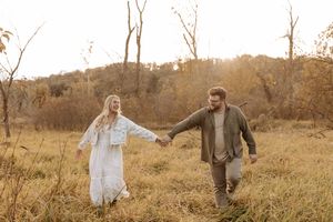 A smiling couple holds hands while walking through a golden autumn field with bare trees and warm sunset light behind them.