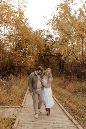 Smiling couple walking arm in arm along a wooden boardwalk through golden autumn trees during a fall engagement photo session.