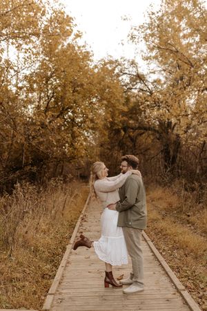 Couple embracing on a wooden boardwalk surrounded by golden autumn trees, the woman in a white skirt lifting her boot playfully.
