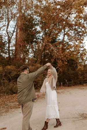 Couple dancing outdoors on a dirt path surrounded by autumn trees, the woman in a white dress and boots twirling under the man's raised arm.