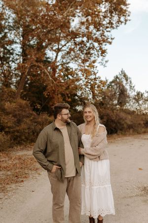 Smiling couple in casual fall outfits stand close on a dirt path surrounded by autumn trees during an outdoor engagement photo session.