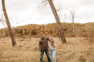 Couple laughing together in autumn field surrounded by golden trees and tall grass on overcast day.