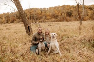 Couple kneeling with two dogs in autumn field with golden trees.